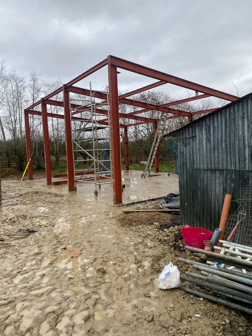 A red steel frame put up on a cleared site next to an old metal barn