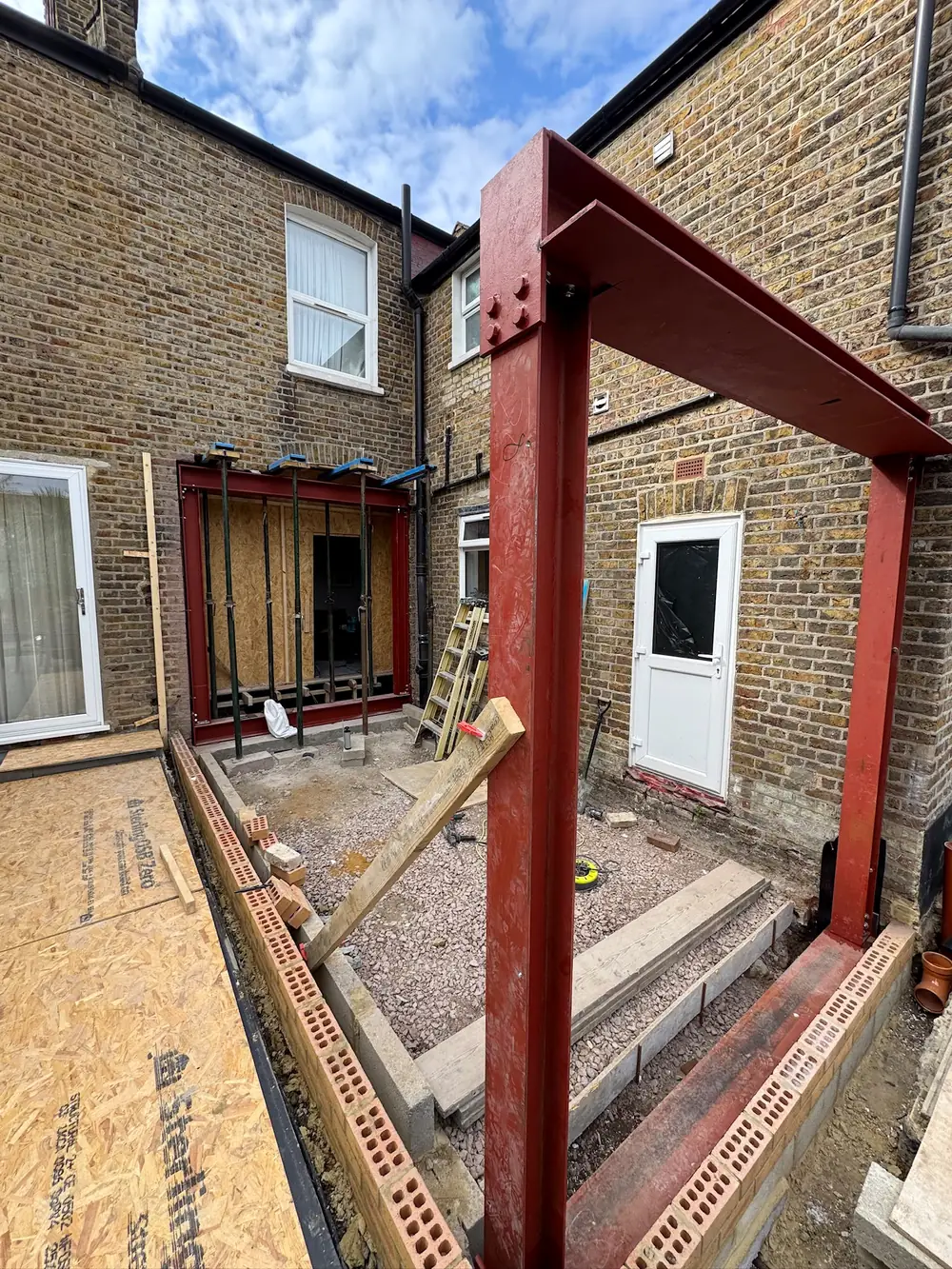A red steel frame standing in the back of a house, bolted to the brickwork, ready for the extension to be built around it