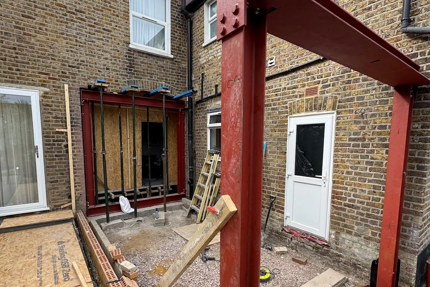 A red steel frame standing in the back of a house, bolted to the brickwork, ready for the extension to be built around it